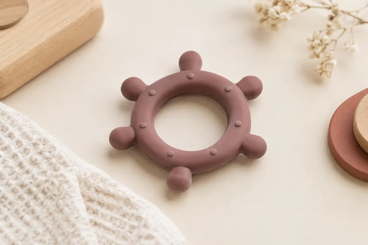 Anneau de dentition rond en silicone présenté sur un fond crème avec du coton gaufré et du bois clair.