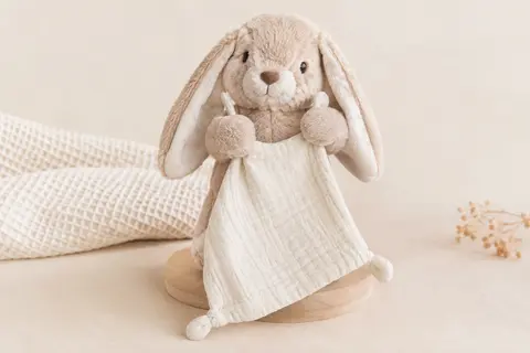 Doudou lapin marionnette en peluche douce tenant un carré de tissu, présenté sur un fond crème lumineux