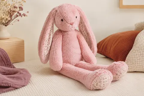 Peluche lapin rose de 40 cm aux longues oreilles et aux longues pattes, posée sur un fond crème près d’un tissu gaufré.