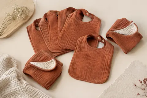Lot de bavoirs bébé en coton terracotta pliés sur un fond crème et du bois clair