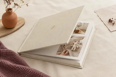 Album de naissance carré à couverture en lin crème, posé ouvert sur un décor clair avec des photos et du papier texturé.