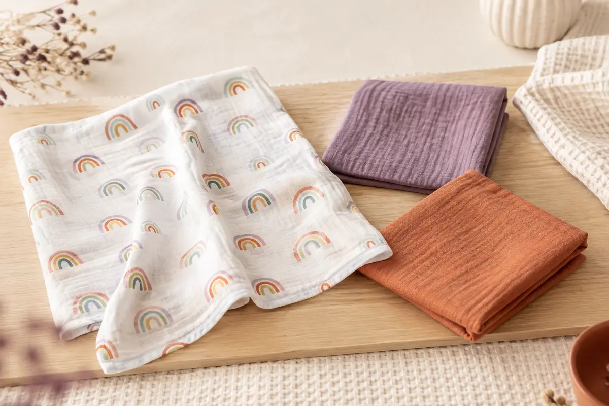 Trois langes bébé en viscose de bambou aux couleurs arc-en-ciel, pliés et dépliés sur un fond crème