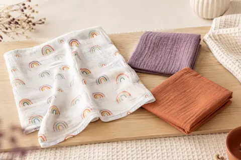 Trois langes bébé en viscose de bambou aux couleurs arc-en-ciel, pliés et dépliés sur un fond crème