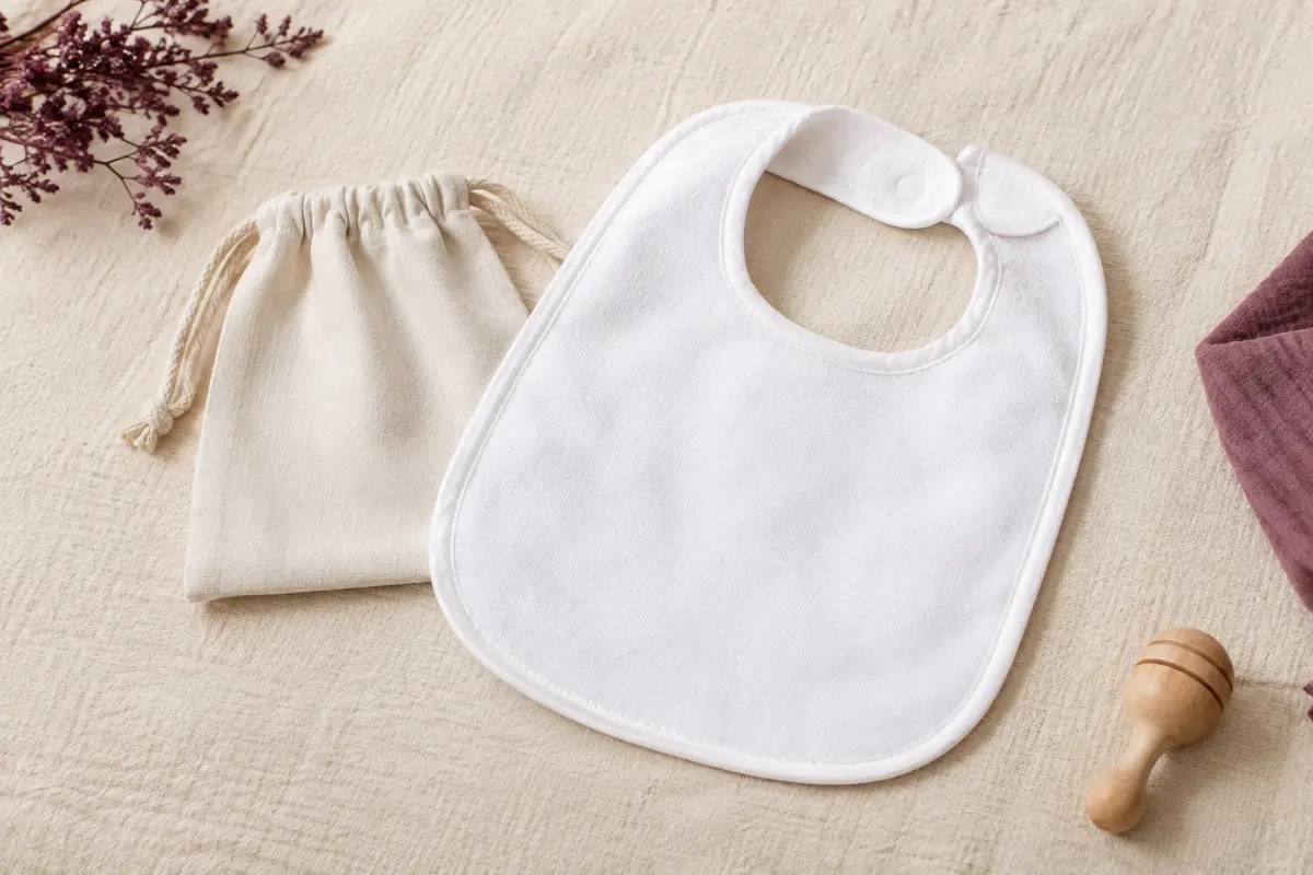 Bavoir bébé blanc en polyester posé avec sa pochette cadeau sur un fond crème lumineux.