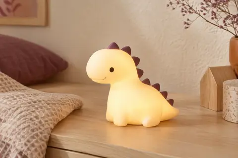 Veilleuse en silicone en forme de dinosaure, allumée sur une surface claire près d’un tissu gaufré.