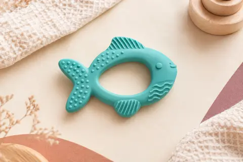 Anneau de dentition Nuby en silicone turquoise, texturé et en forme de poisson, présenté sur un fond crème