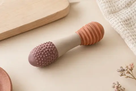 Jouet de dentition tubulaire en silicone souple aux tons doux, posé sur un fond crème près de bois clair.