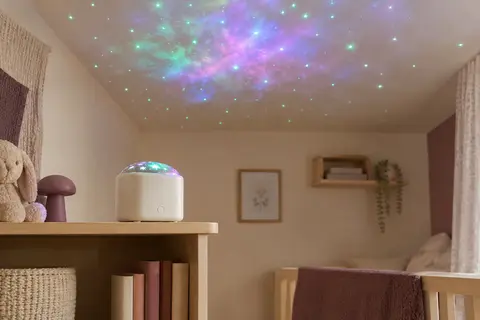Veilleuse-projecteur blanche posée sur une étagère, avec une nébuleuse colorée et des étoiles projetées au plafond.