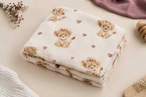 Couverture bébé en peluche imprimée d’un lionceau et de petits cœurs, présentée sur un fond crème.