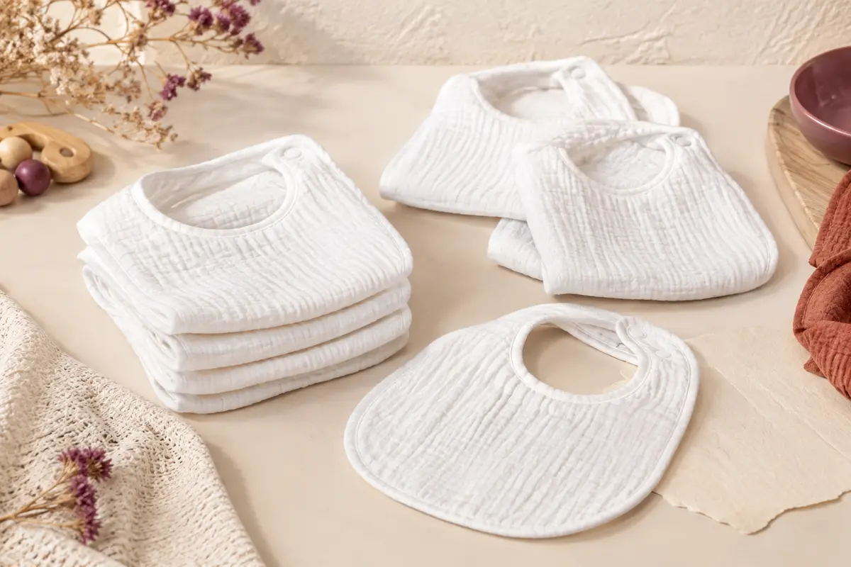 Sept bavoirs bébé blancs en tissu double épaisseur, disposés sur un fond crème et du bois clair.