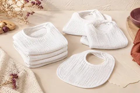 Sept bavoirs bébé blancs en tissu double épaisseur, disposés sur un fond crème et du bois clair.