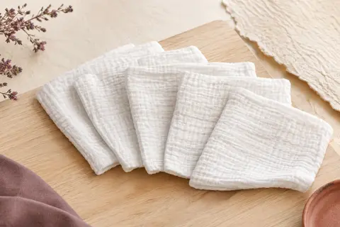 Cinq langes blancs en mousseline de coton pliés sur un fond crème lumineux