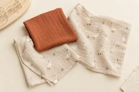 Trois langes en mousseline de coton, un uni terracotta et deux beiges à motif de petites oies.